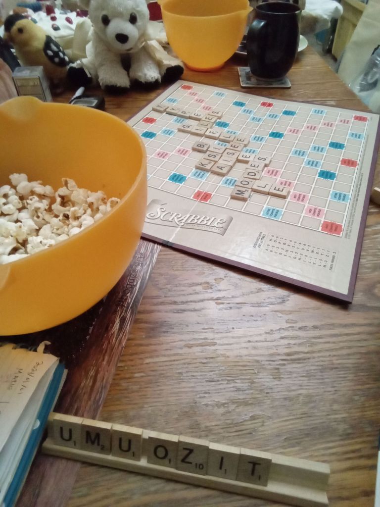 Un plateau de Scrabble avec quelques mots, en avant-plan, mes lettres U, M, U, O, Z, I, T, et de côté un grand bol rempli de popcorn.