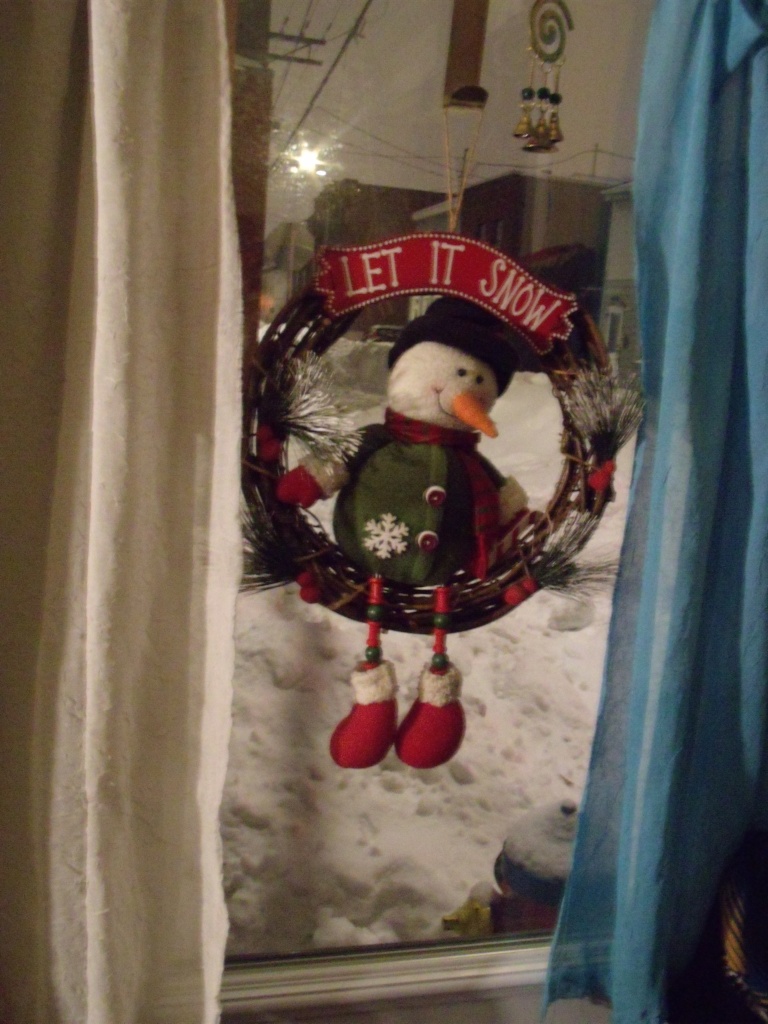 Une couronne faite de vigne sur laquelle un bonhomme de neige en peluche est assis est suspendu dans la fenêtre, dehors, une épaisse couverture de neige recouvre la rue.